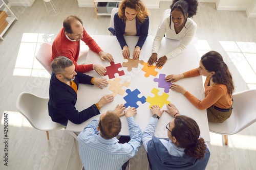 Naklejka Teamwork concept. Diverse business team meeting around office table and trying to fit jigsaw puzzle together. Young and senior people working on project, looking for solution and suggesting ideas