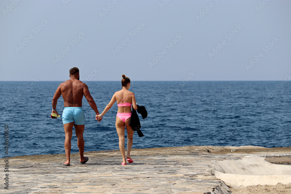 Couple walking on a stone beach on sea background. Muscular tanned man in blue trunks and girl with white skin in pink bikini together, romantic holiday and vacation