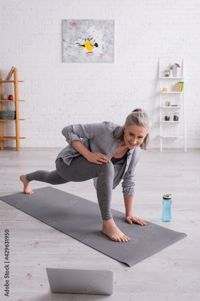 © LIGHTFIELD STUDIOS - happy mature woman with grey hair practicing yoga on mat near sports bottle while watching tutorial on laptop