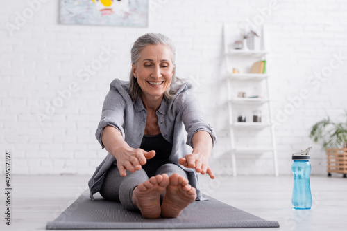 happy mature woman with grey hair stretching on yoga mat near sports bottle
