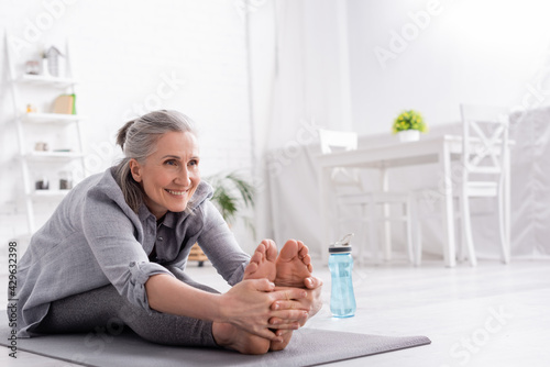 cheerful mature woman with grey hair stretching on yoga mat near sports bottle photo