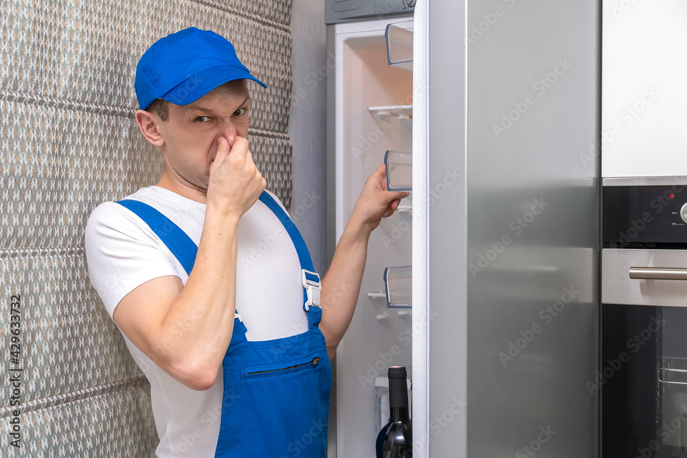 Uniformed repairman covers his nose from the unpleasant smell, looks at the camera and points to the open refrigerator in the kitchen Uniformed repairman covers his nose from the unpleasant smell, looks at the camera and points to the open refrigerator in the kitchen