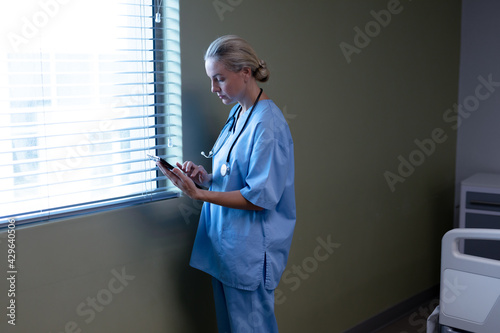 Serious caucasian female doctor in hospital wearing scrubs and stethoscope using tablet