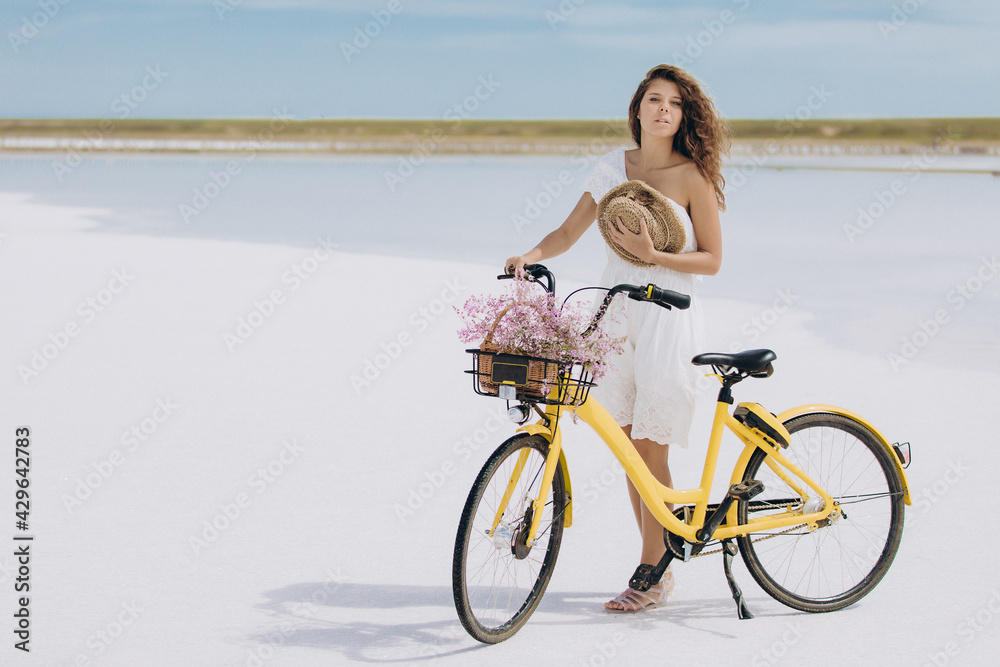 Fototapeta premium Young beautiful girl with long hair in a white dress near a yellow bicycle with flowers on a white and pink salt lake