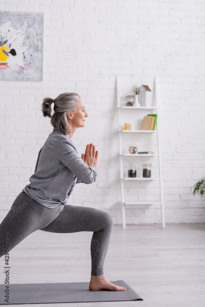© LIGHTFIELD STUDIOS - middle aged woman in warrior with praying hands pose practicing yoga on mat at home