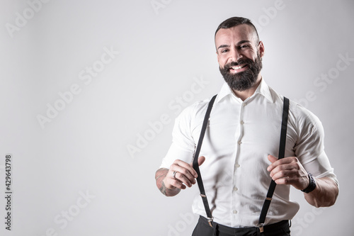 Uomo moro con la barba con camicia bianca tiene le bretelle nere con espressione divertita, isolato su sfondo  photo