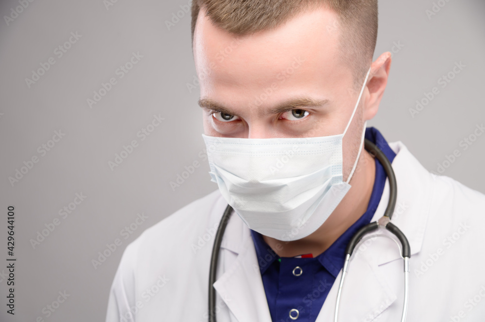A young Caucasian doctor wearing a mask looks ominously into the camera from under his brow. Medical conspiracy concept against humanity A young Caucasian doctor wearing a mask looks ominously into the camera from under his brow. Medical conspiracy concept against humanity