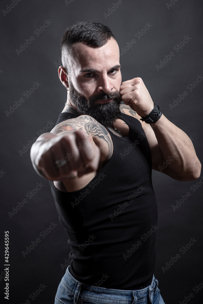 Uomo moro muscoloso, tatuato con la barba, stai un posizione di combattimento pronto all'attacco , isolato su sfondo nero