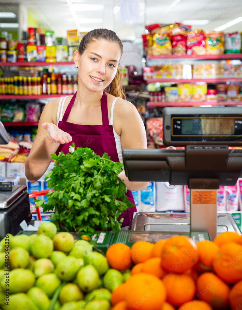 Positive young saleswoman working in the store shows fresh greens, standing near the scales and counter