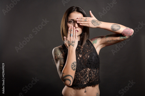 bellissima ragazza  mora con i tatuaggi scopre la bocca l'occhio con l'altra mano, isolata su sfondo nero photo