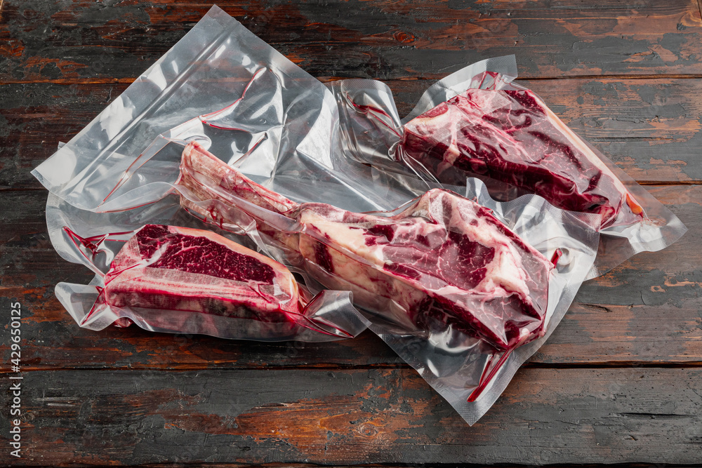 Dry aged steak in a vacuum marbled beef meat pack, tomahawk, t bone and club steak cuts, on old dark  wooden table background