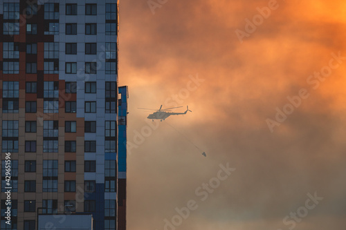 Fire fighting helicopter silhouette with bambi bucket for сarrying water to put out a massive building city fire, process of put out a large blaze bush fire wildifre, aerial firefighting with chopper