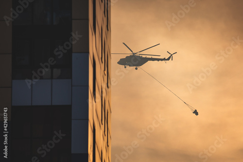 Fire fighting helicopter silhouette with bambi bucket for сarrying water to put out a massive building city fire, process of put out a large blaze bush fire wildifre, aerial firefighting with chopper photo