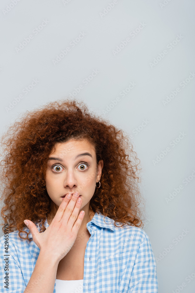 shocked woman with wavy hair covering mouth with hand isolated on grey shocked woman with wavy hair covering mouth with hand isolated on grey
