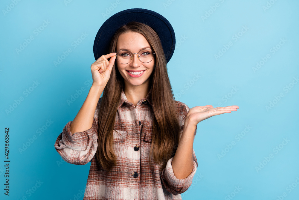 Photo of happy positive smiling cheerful woman in glasses and hat advertising product isolated on blue color background Photo of happy positive smiling cheerful woman in glasses and hat advertising product isolated on blue color background