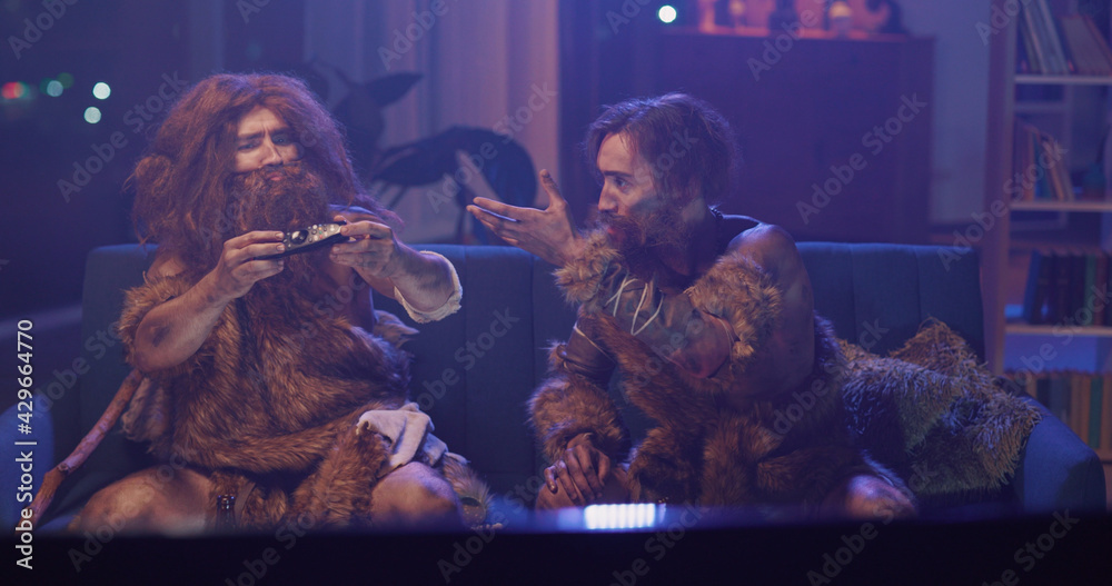 Fototapeta premium Funny Couple of Ancient Neanderthals Sitting in Living Room Interacting Television Watching Soccer Prehistoric Game in Modern Apartment. Evolution Concept.