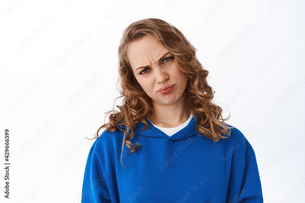 Skeptical and moody blond girl grimacing, frowning displeased, looking disappointed and upset, standing over white background Skeptical and moody blond girl grimacing, frowning displeased, looking disappointed and upset, standing over white background