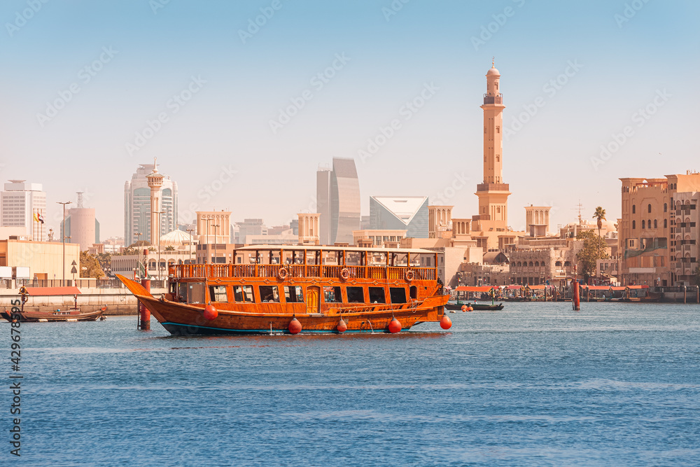An authentic wooden cruise ship cruises and transports tourists along the Dubai Creek Canal with skyscrapers and minarets of mosques in the background. Travel and sightseeing - obrazy, fototapety, plakaty 