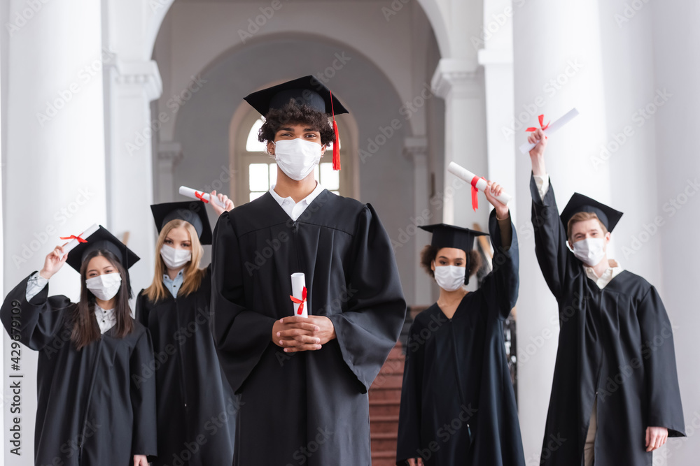 African american graduate in medical mask holding diploma near friends on blurred background
