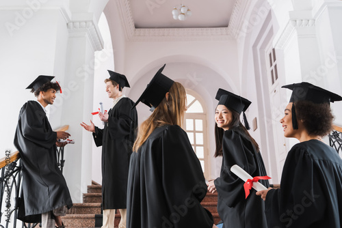 Interracial graduates in academic dresses and caps talking in university