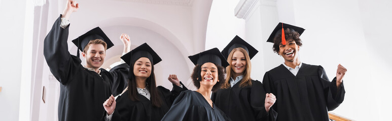 Cheerful multiethnic students in academic gowns showing yes gesture, banner