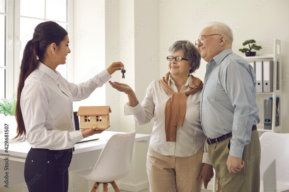 Fototapeta premium Female real estate agent give key from new home to excited elderly wife and husband client couple. Woman realtor or broker congratulate happy mature spouses with house purchase. Ownership concept