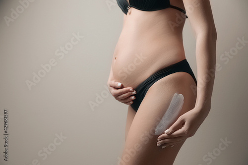 Pregnant woman applying sun protection cream on her leg against beige background, closeup. Space for text