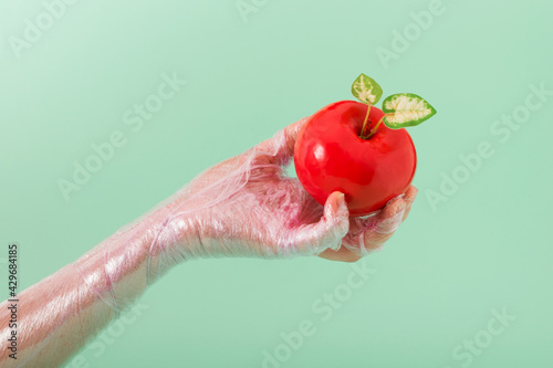 Plastic apple in hand on pastel green background. Minimalistic summer and GMO food concept.