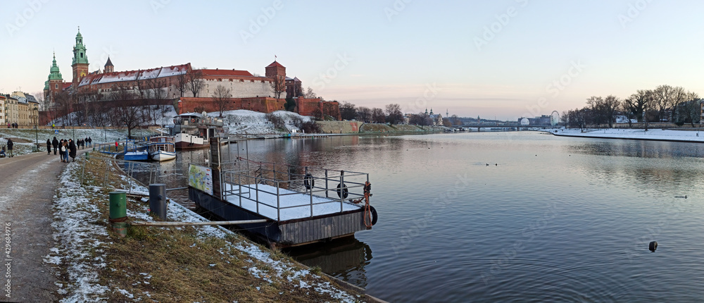 Krakow, Poland : Wide angle shot of a wawel castle and people taking a walk on a street next to vistula river during some snow on the ground.