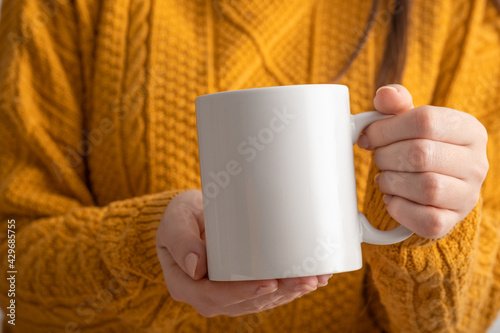 Female hands hold mock up white empty mug, cup for your design and logo close-up. Woman in yellow knitted sweater autumn winter. Blank template for promotional text message or promotional content.