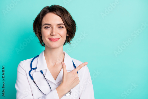 Photo of young woman doctor happy positive smile indicate finger empty space ad advice clinic isolated over teal color background photo