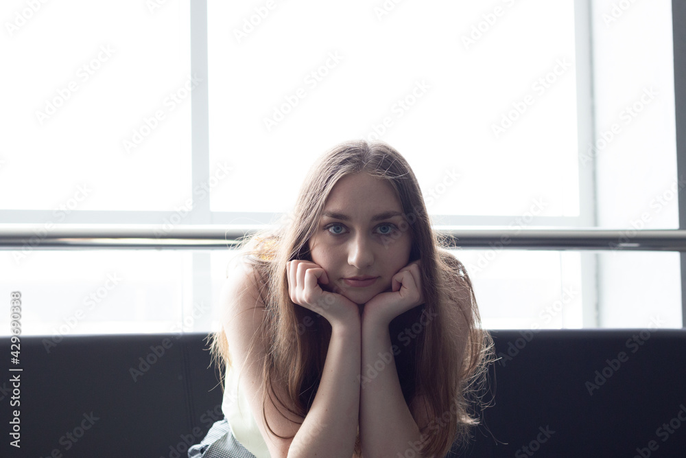 portrait of a beautiful young long blond hair and blue eyes looking woman sitting on sofa near window and holding head on hands portrait of a beautiful young long blond hair and blue eyes looking woman sitting on sofa near window and holding head on hands