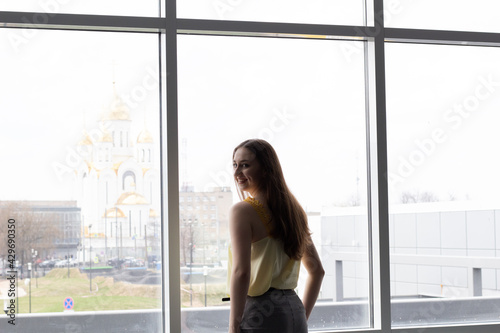 portrait of a beautiful young long blond hair and blue eyes woman in yellow blouse standing near window with city view on church Free Download portrait of a beautiful young long blond hair and blue eyes woman in yellow blouse standing near window with city view on church - GFXLoad