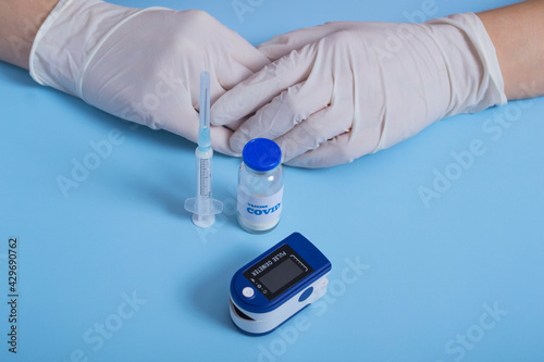 Medical concept. On a blue background doctor's hands in white gloves,pulse oximeter, coronavirus vaccine, syringe. virus protection.Concept: prevention of coronavirus