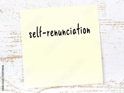 Yellow sticky note on wooden wall with handwritten word self-renunciation photo