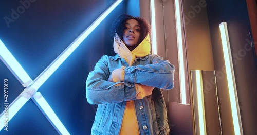 Wide angle portrait of stylish urban african american black woman in yellow hoodie and jeans jacket standing by wall in night disco club backlit with purple and blue neon light stripes. Urban style photo