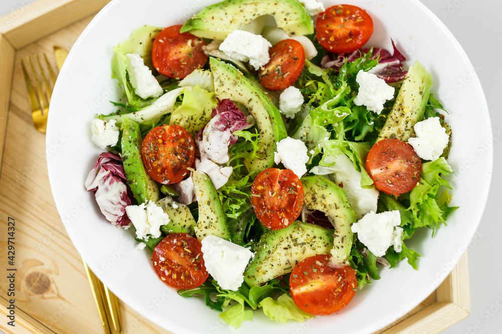 Fresh green salad with avocado, tomatoes, goat cheese in big white bowl on wooden tray. Vegan salad with green mix leaves and vegetables. Top view on gray table. Healthy vegetarian food. Close up Fresh green salad with avocado, tomatoes, goat cheese in big white bowl on wooden tray. Vegan salad with green mix leaves and vegetables. Top view on gray table. Healthy vegetarian food. Close up