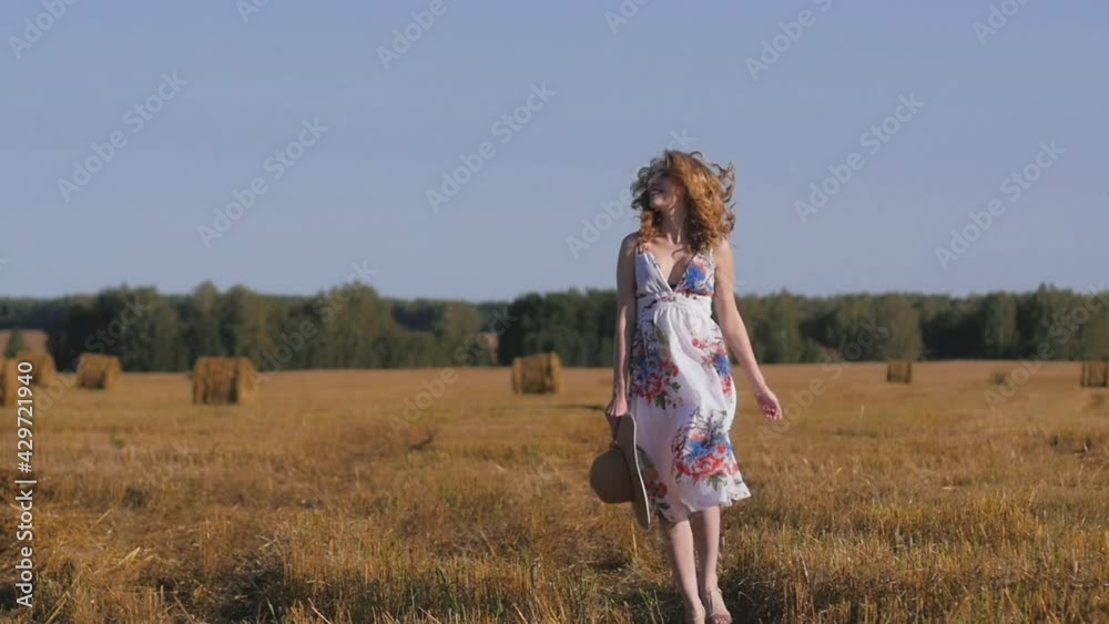 beautiful red-haired woman in a white dress and hat cheerfully walks on the background of fields and haystacks. Young woman at sunset walks towards the camera, bursts her hair and smiles beautiful red-haired woman in a white dress and hat cheerfully walks on the background of fields and haystacks. Young woman at sunset walks towards the camera, bursts her hair and smiles