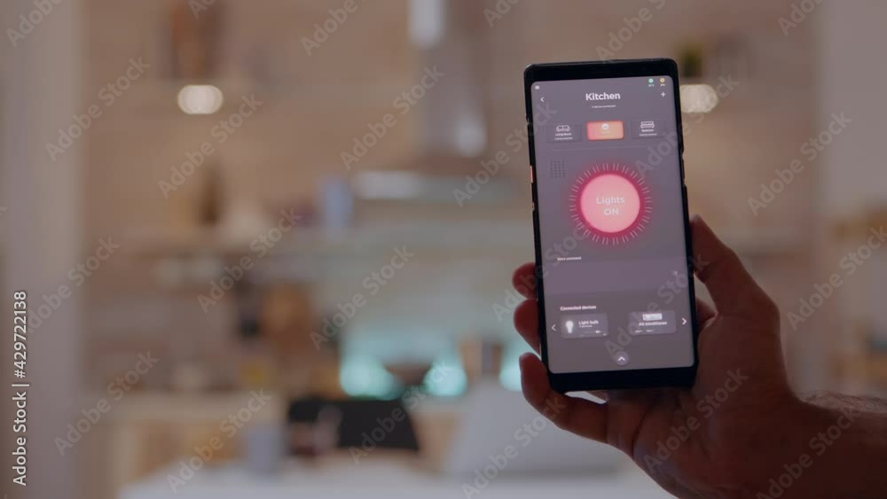 Close up of phone with high tech software lighting controller turning on the light before man working on laptop. Person tapping on screen for switching on the bulb in house with automation system