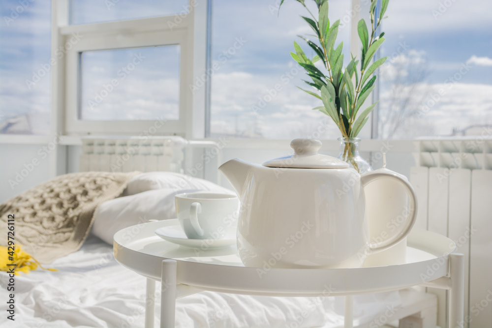 White teapot with tea, white cup stand on a white table in the morning in the bedroom