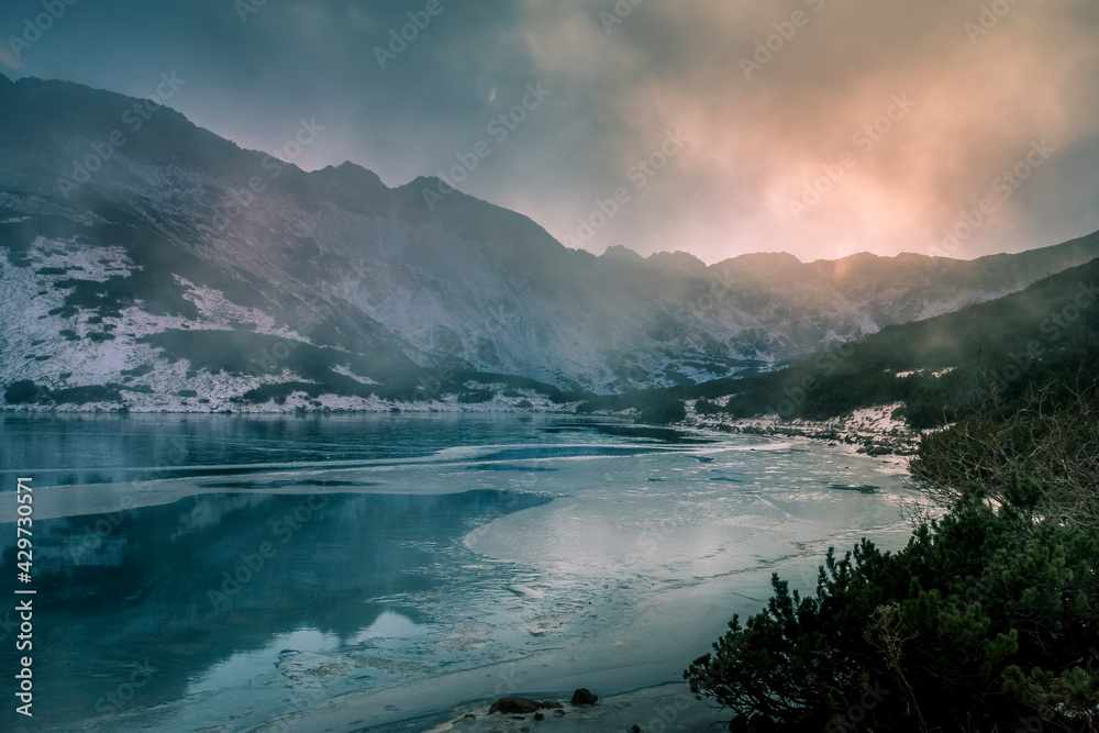 Freezing lake and dramatic sky in High Tatra Mountains, Poland. Ice is covering the surface of water, clouds are rising over the valley. Selective focus on the rocks, blurred background.