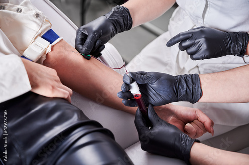 Close up of doctor hand in black gloves holding test tube with blood while assistant inserting needle into patient arm. Medical laboratory scientists collecting blood sample from young woman.