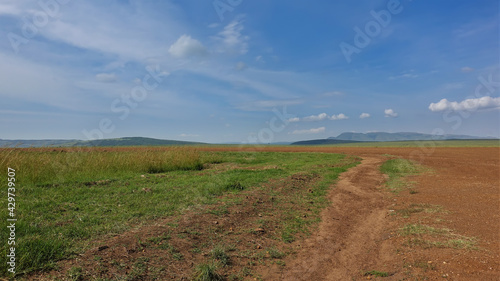 Landscape of the African savannah. The dirt road runs along the red earth. Lush green grass grows on the roadside. Silhouettes of mountains against the blue sky. Kenya. Masai Mara © Вера 