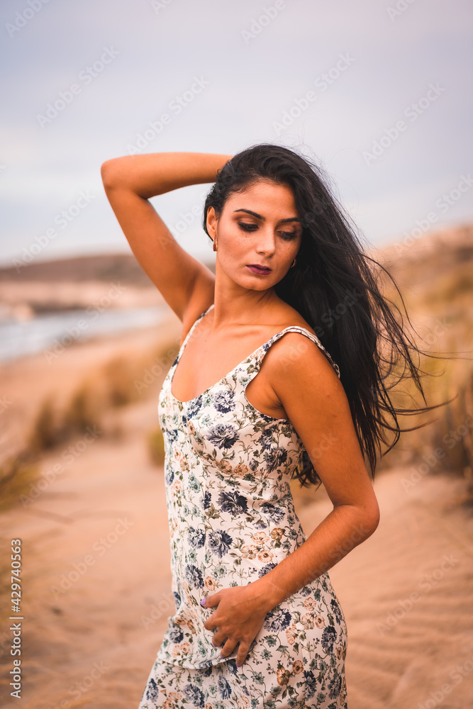 A Caucasian brunette in a white flower dress, in the sand on the beach, vertical photo A Caucasian brunette in a white flower dress, in the sand on the beach, vertical photo