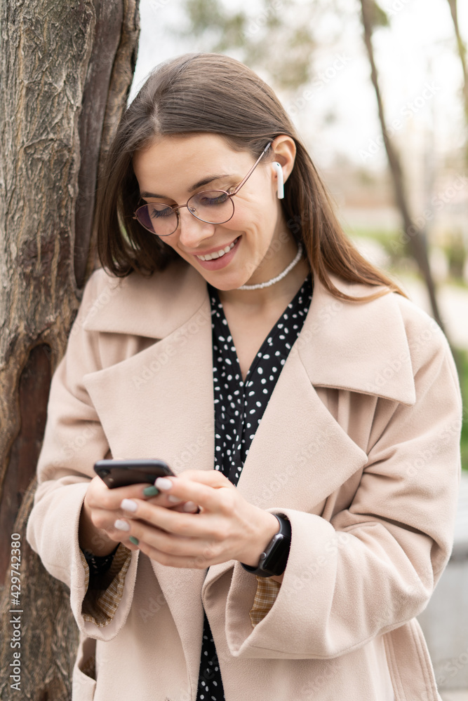 Young business woman texting on her mobile phone in the park wearing eyeglasses and earphones with a smile, vertical casual attire Young business woman texting on her mobile phone in the park wearing eyeglasses and earphones with a smile, vertical casual attire