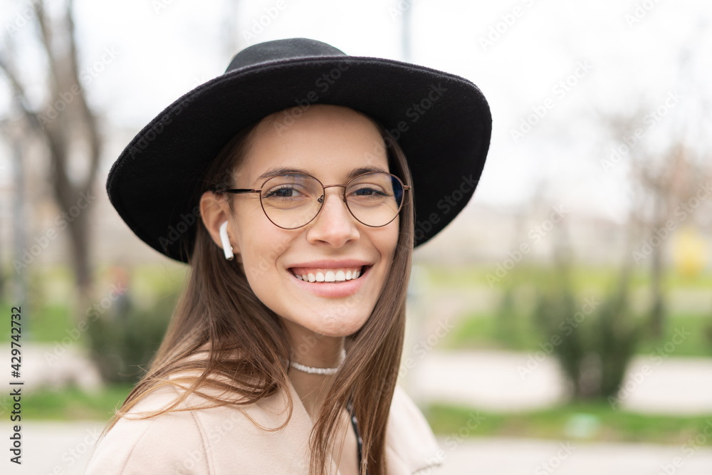 Close up portrait of a smiling woman wearing a hat and eyeglasses outdoors Wirth an earphones, looking in the camera  