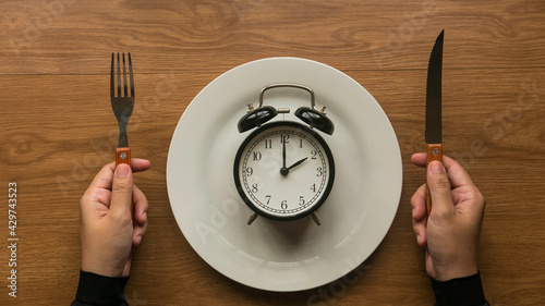Concept of time and eating with a hand holding fork and knife and a vintage alarm clock on a white plate.