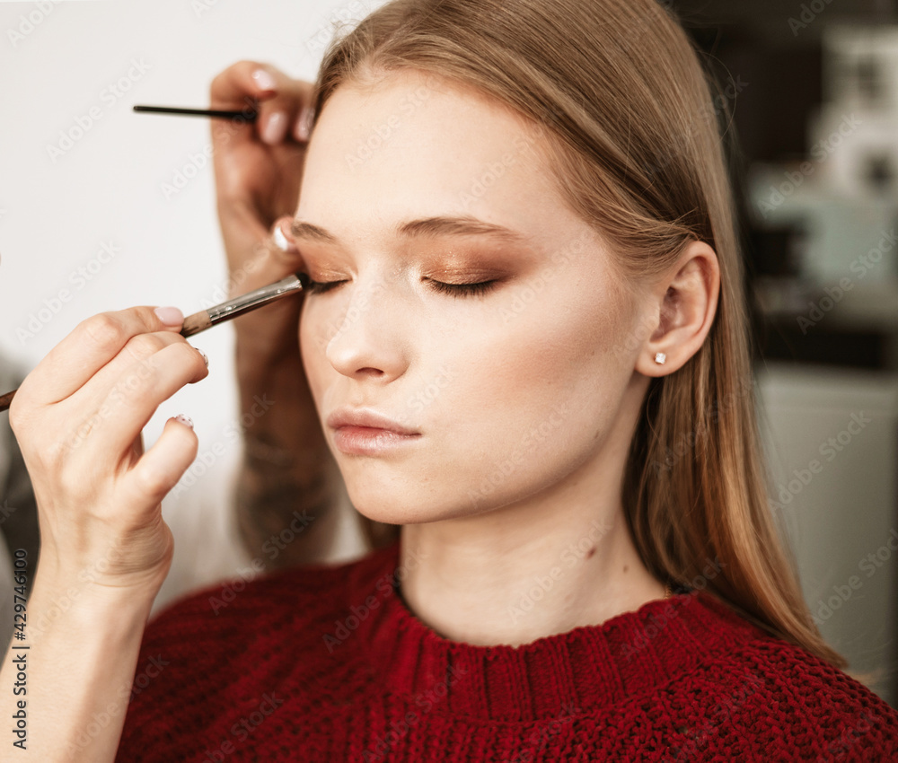 Obraz premium makeup artist applies eye shadow to the eyelids on a beautiful young woman blonde model, face make up concept