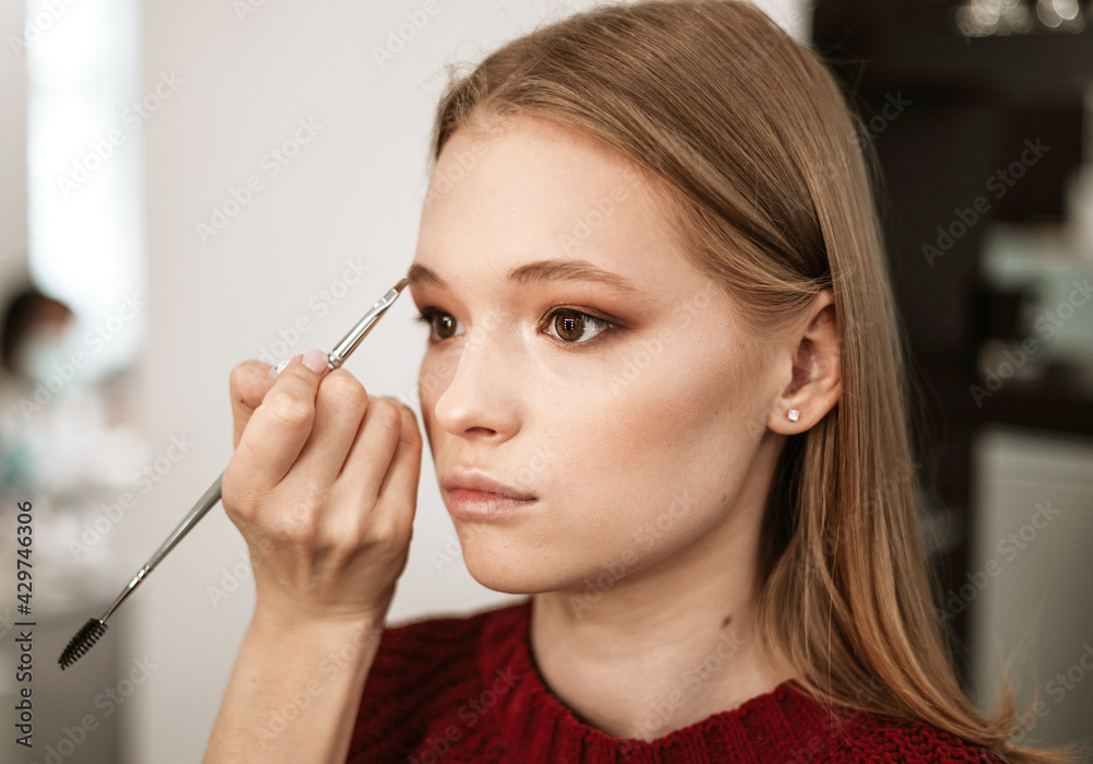 makeup artist applies eyebrow shadow on a beautiful young woman blonde model, face make up concept