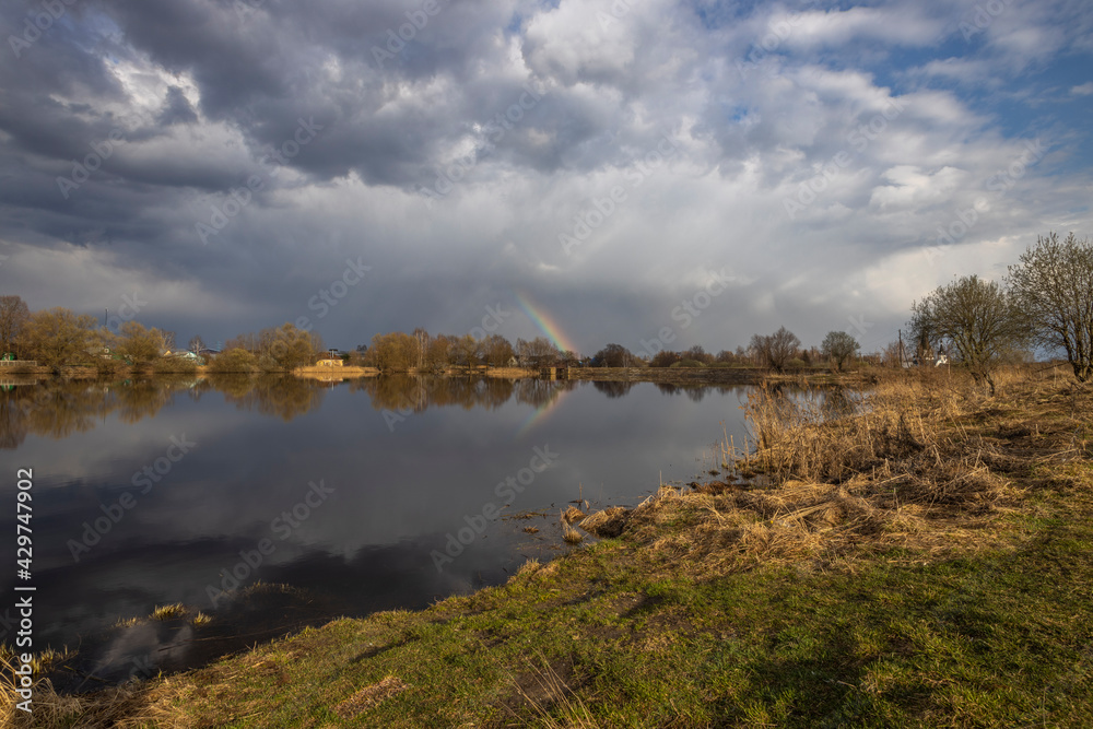 On the horizon is a village by the river. The clouds are reflected in the water. Early spring by the pond. A fragment of a rainbow at the horizon.